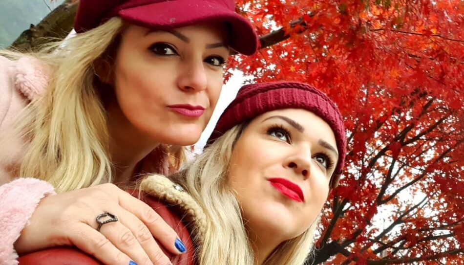 duas mulheres loiras de gorro vermelho em frente a uma árvore de folhas vermelhas