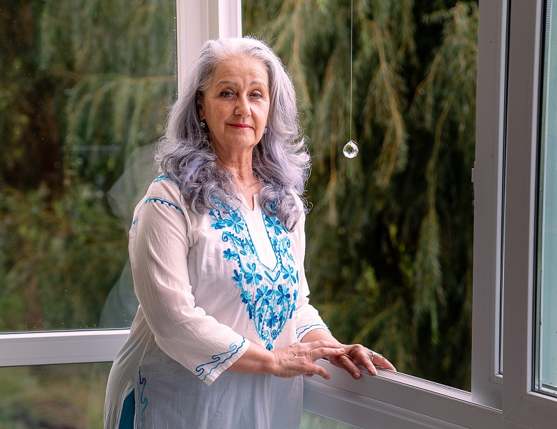 autora do livro mulher com cabelos longos grisalhos perto de uma janela aberta com árvores ao fundo